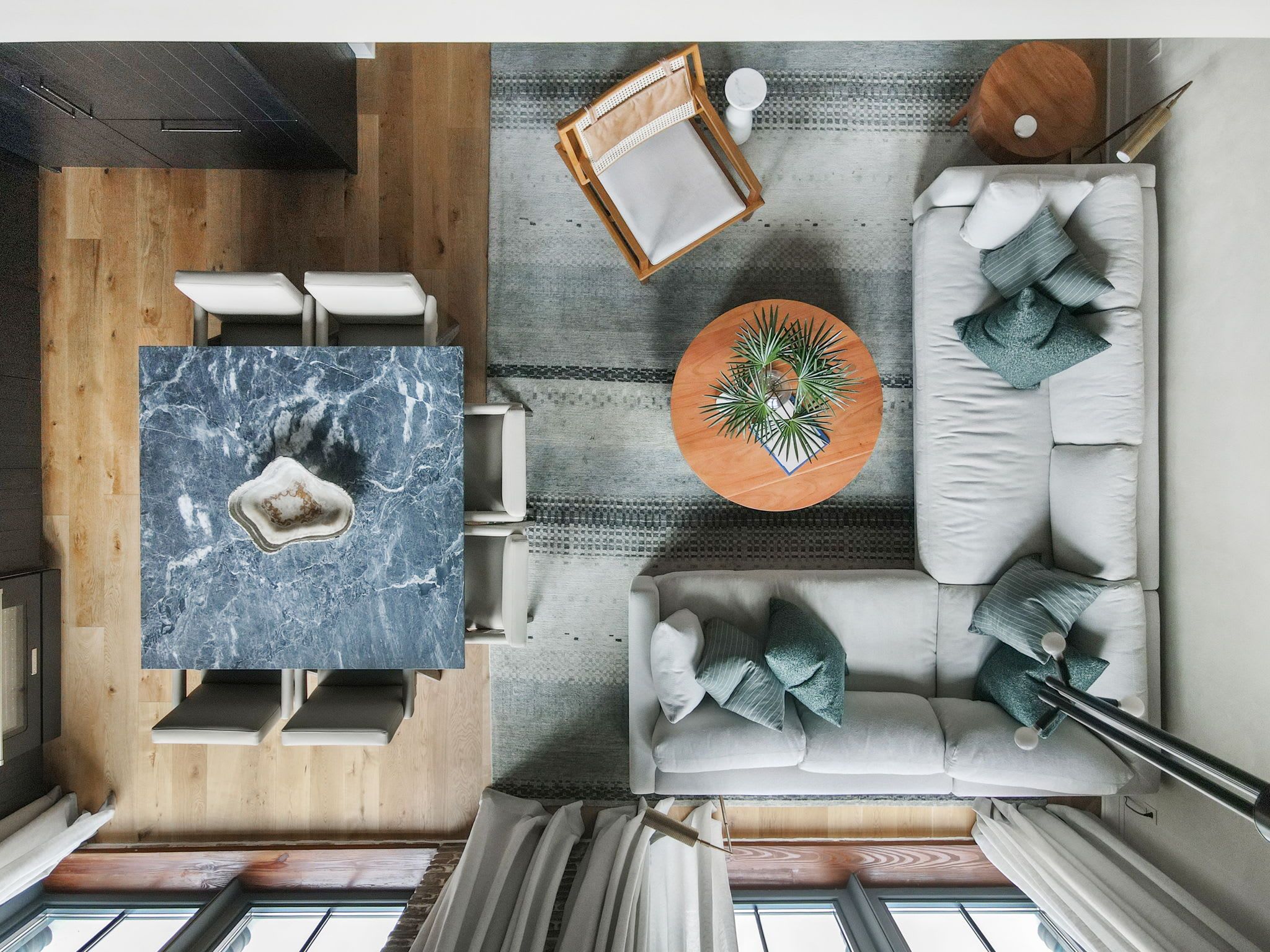 Una sala moderna con una mesa de centro de mármol, una mesa redonda de madera con una planta, un sofá blanco con cojines y vistas al cielo a través de una ventana.