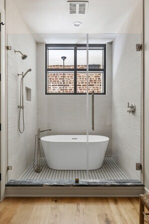 Un baño moderno con una bañera independiente, ducha con mampara de vidrio y vistas a un muro de ladrillo a través de una ventana.