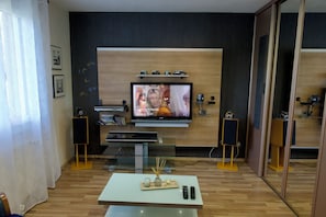 Ein modernes Wohnzimmer mit einem an einer Holzwand montierten Flachbildfernseher, einem Glastisch mit Kerzen und einem Bücherregal.