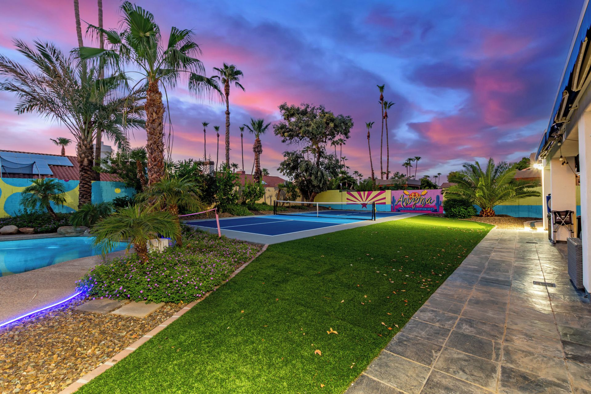 A tennis court with a net, surrounded by palm trees and a colorful mural, under a twilight sky.
