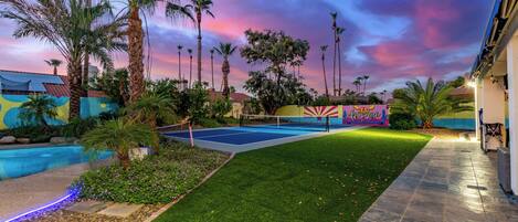 A tennis court with a net, surrounded by palm trees and a colorful mural, under a twilight sky.