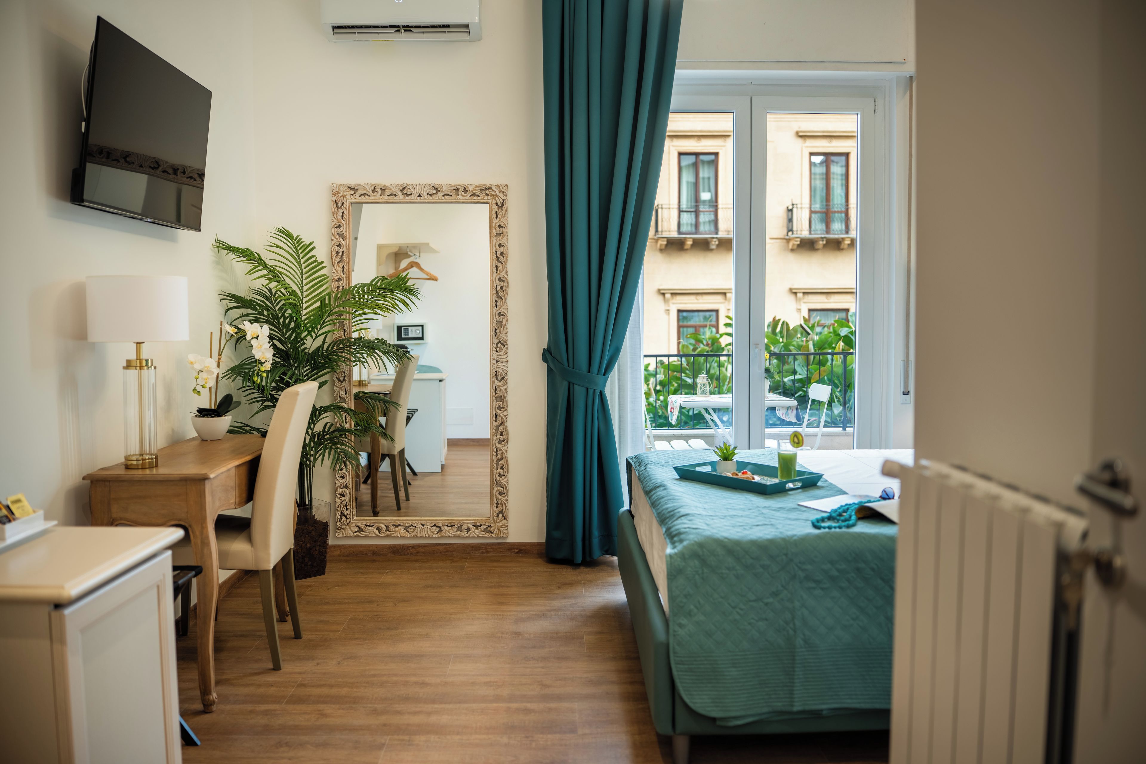 Habitación de hotel con un escritorio de madera, una silla, un espejo, una cama con colcha verde y vistas a edificios a través de la ventana.