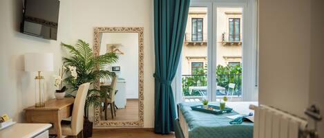 Habitación de hotel con un escritorio de madera, una silla, un espejo, una cama con colcha verde y vistas a edificios a través de la ventana.