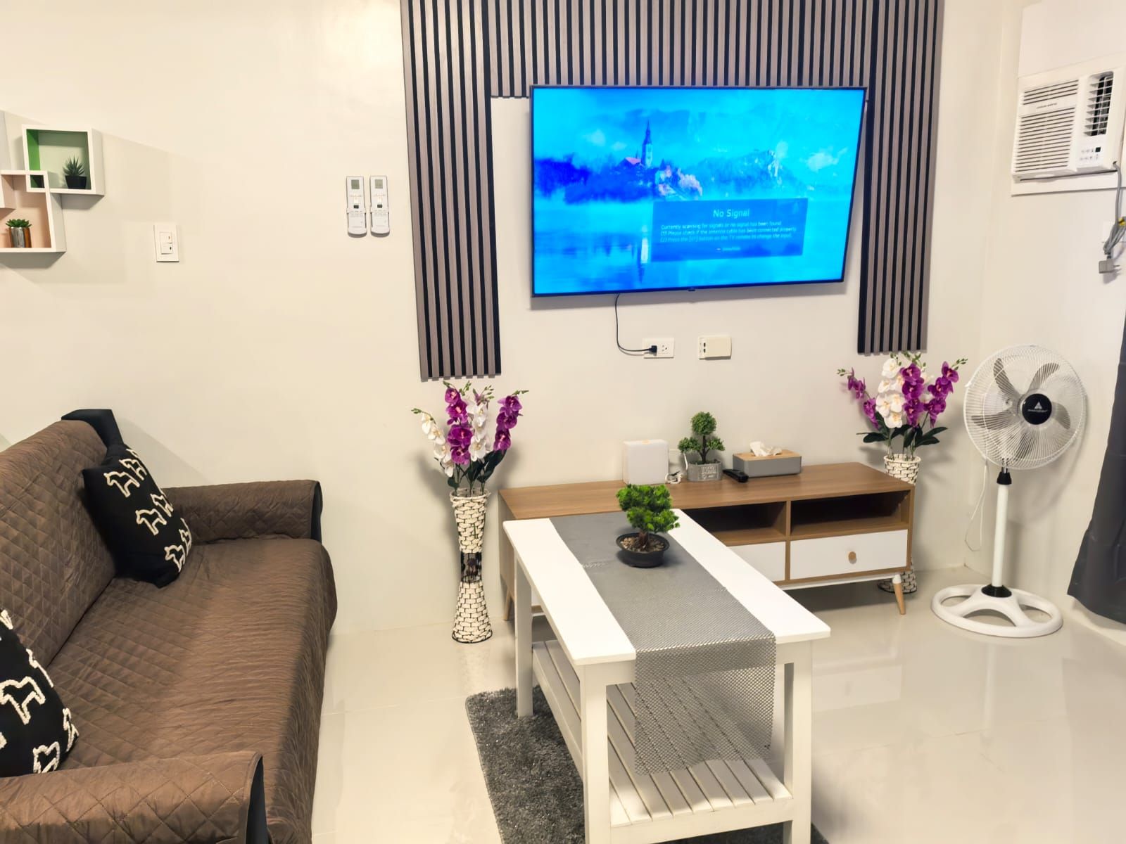 Ein modernes Wohnzimmer mit einem an der Wand befestigten Flachbildfernseher, einem braunen Sofa, einem weißen Couchtisch und zwei Zimmerpflanzen.