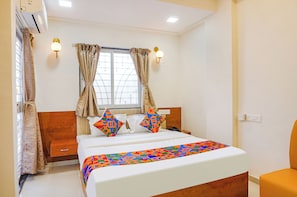 A hotel room with a bed, a bedside table, a lamp, and a window with curtains. - RISING SURYA HOTEL (Pune)
