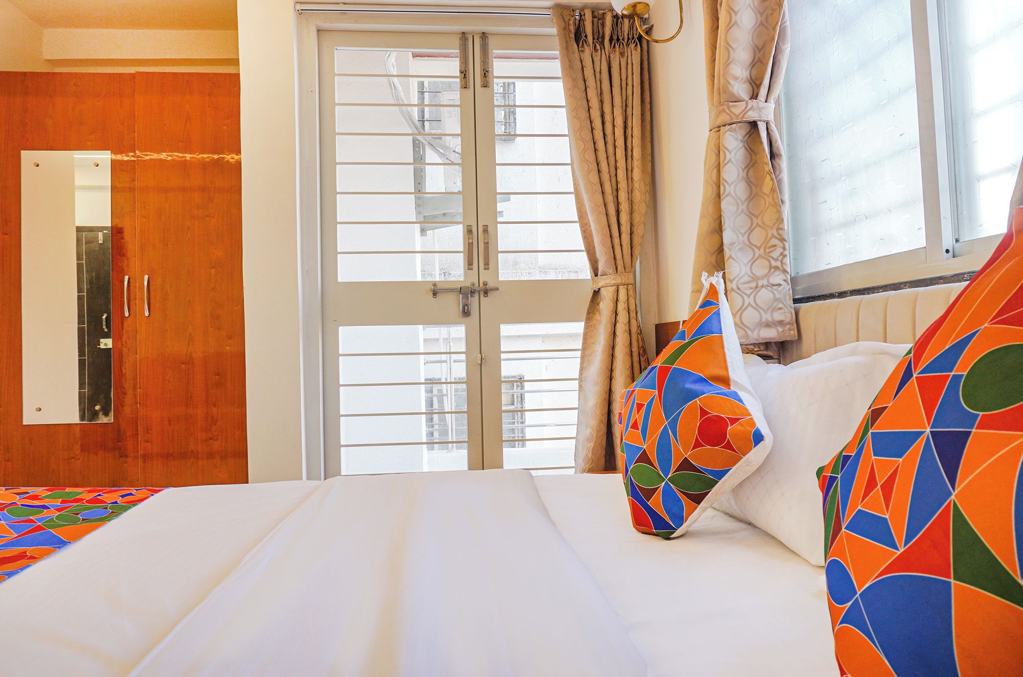Una habitación de hotel con cama, ventana con cortinas, puerta de madera y espejo adosado a la pared.