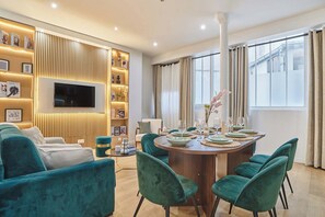 Ein modernes Wohnzimmer mit einem runden Esstisch, stuhlartigen Sitzgelegenheiten in Türkis, einem Flachbildfernseher, der an einer Holzwand angebracht ist, und einem Bücherregal mit Zeitschriften und Dekorationsobjekten.