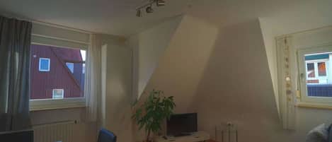 Une pièce avec un plafond en pente, un téléviseur posé sur un meuble, une plante en pot et une fenêtre avec des rideaux.