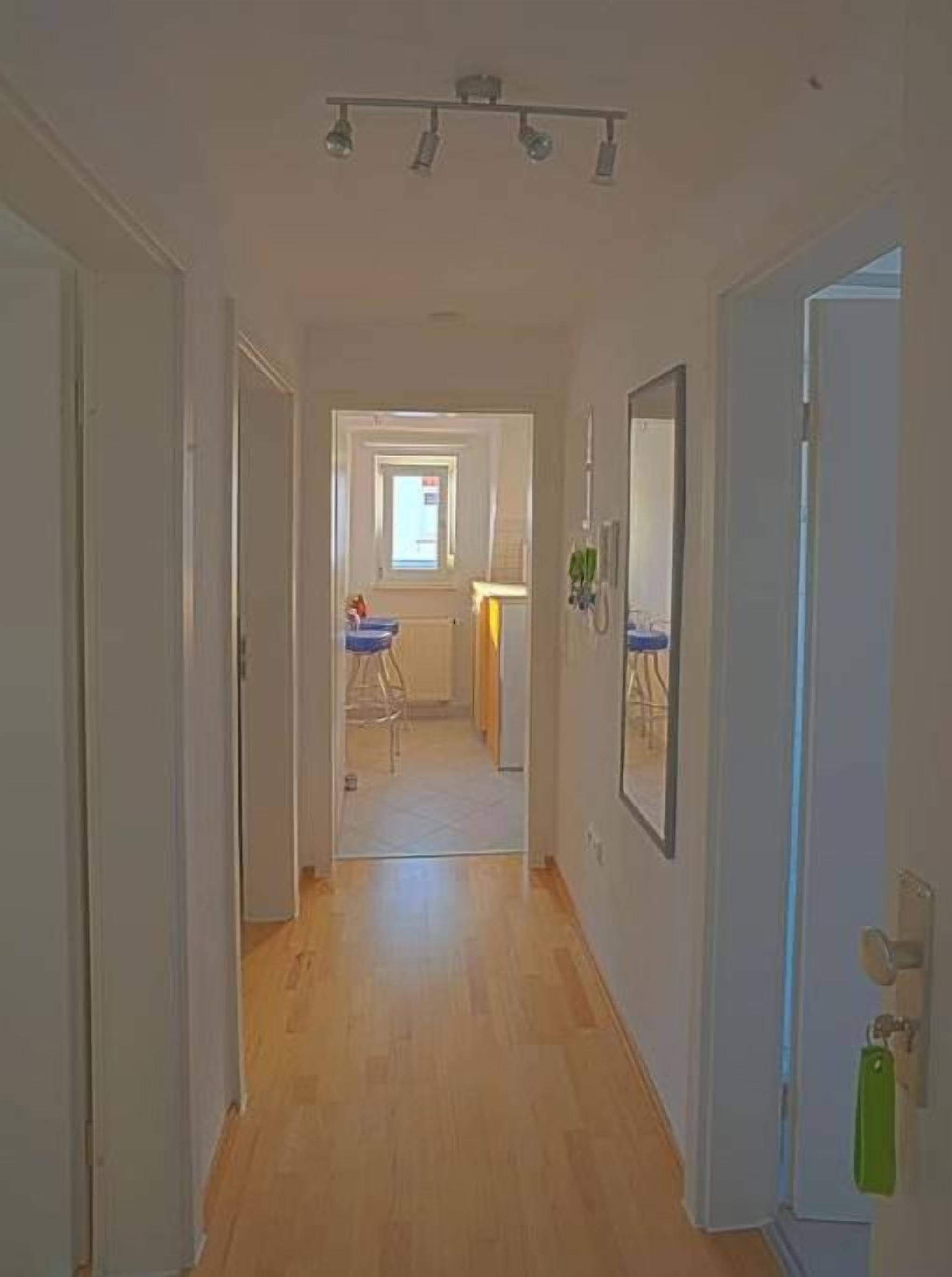 Um corredor com piso de madeira, uma porta à direita e uma janela no final.