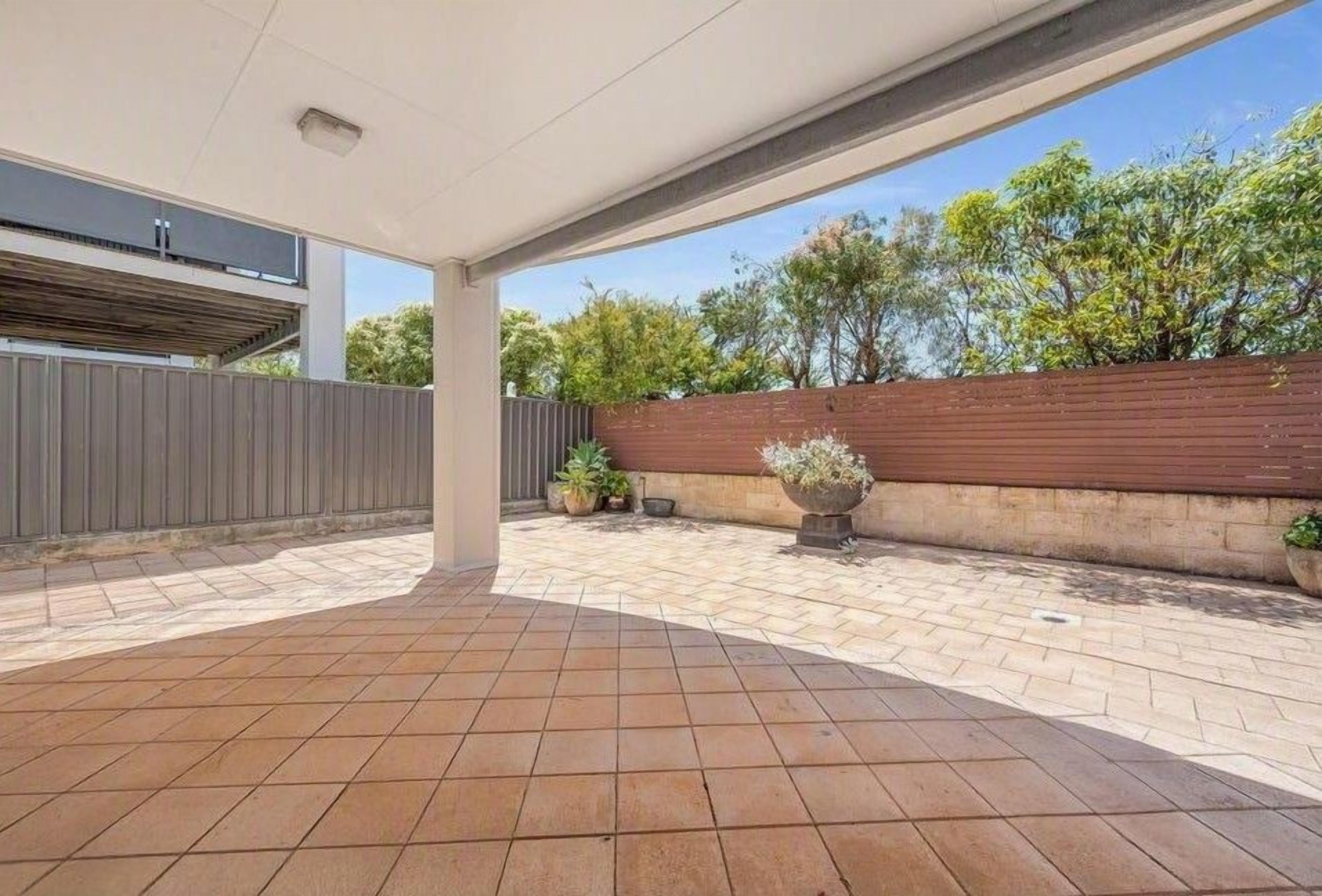 A covered outdoor patio area with tiled flooring, a wooden fence, and potted plants.