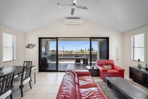 A spacious living room with a red leather sofa, a dining table with chairs, a red armchair, and a ceiling fan. - Coastal luxury home with breathtaking ocean and harbour views (Robe)