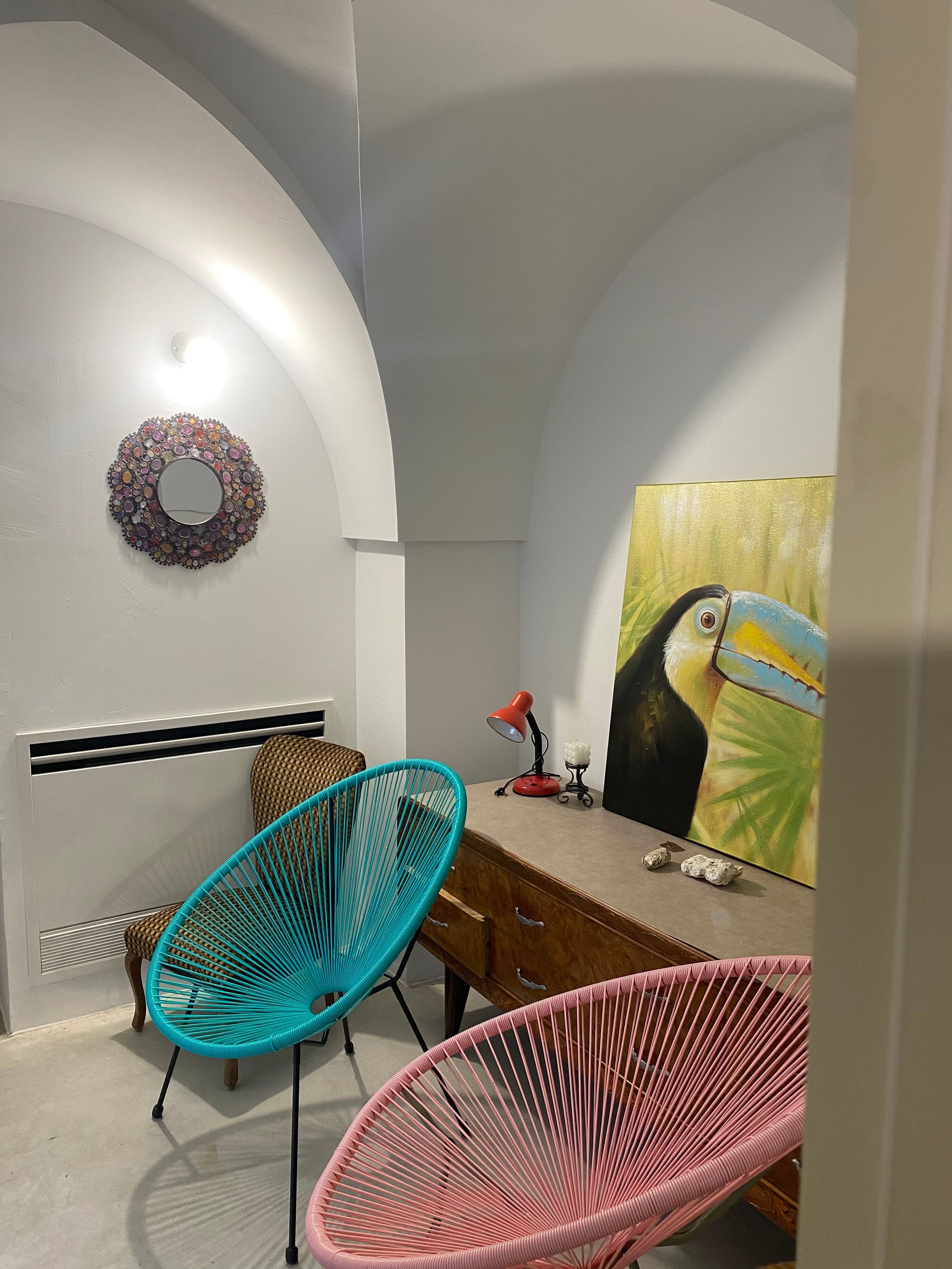 Une pièce avec un plafond incurvé, un miroir rond, un tableau représentant un oiseau, deux chaises, un bureau et une lampe rouge.