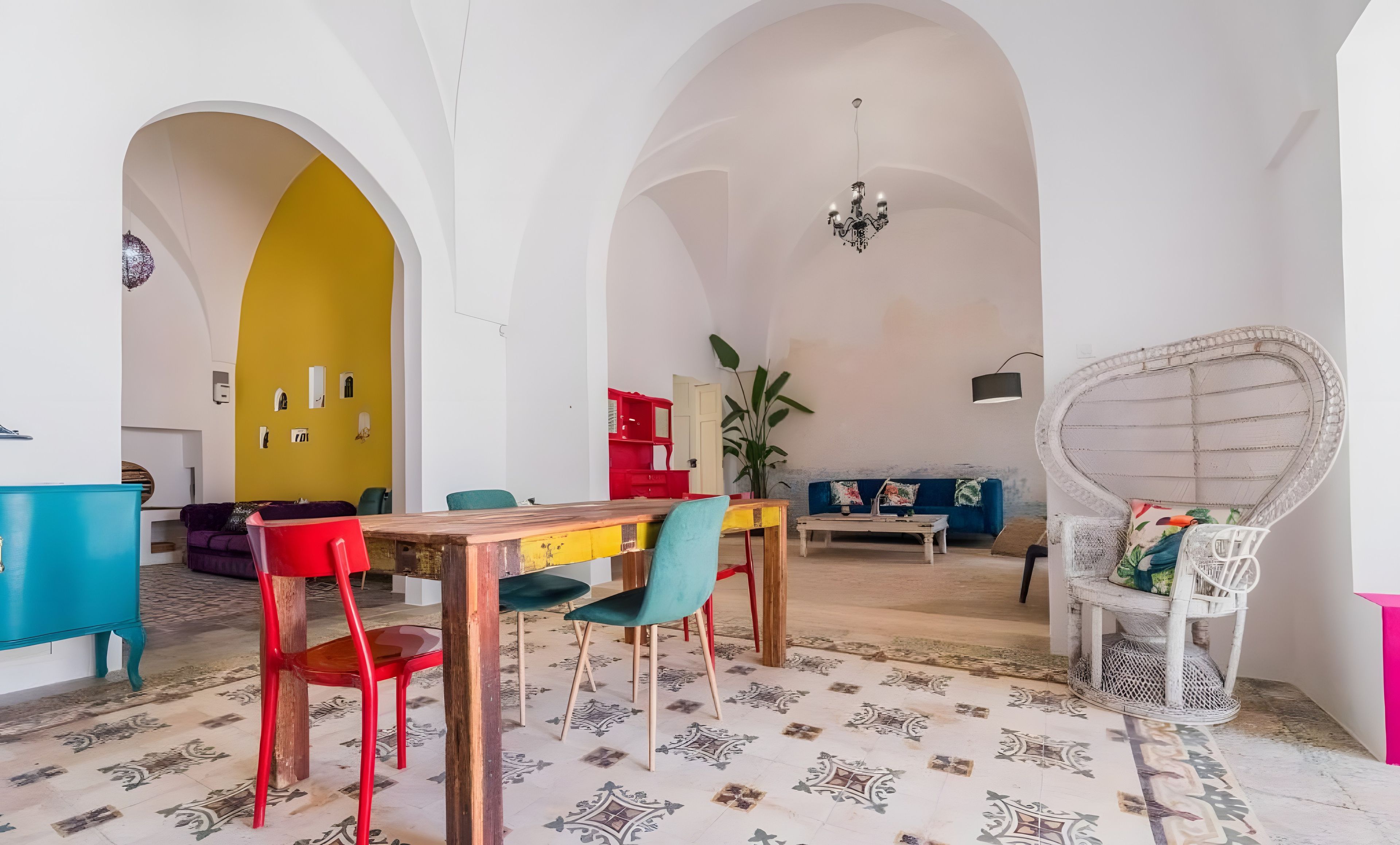 Un área de comedor con una mesa de madera, sillas coloridas y un sofá azul en una habitación con arcos blancos y una pared amarilla.