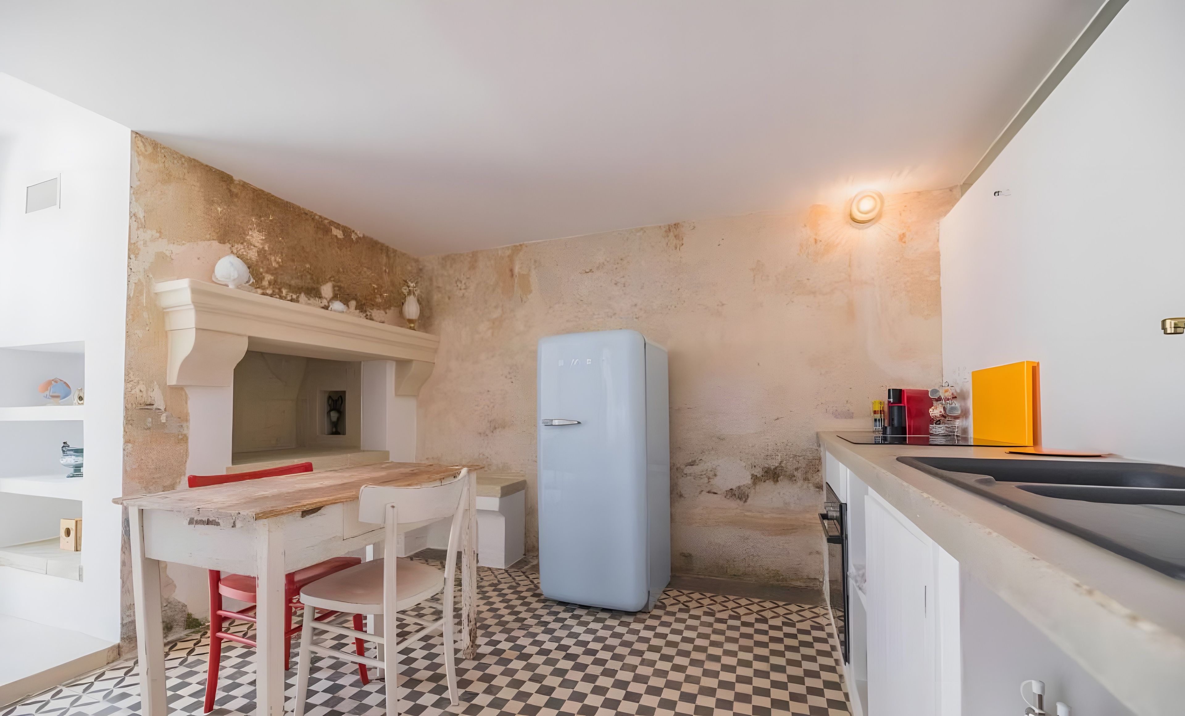 A compact kitchen with a checkered floor, a white table with red chairs, a blue refrigerator, and a small sink.