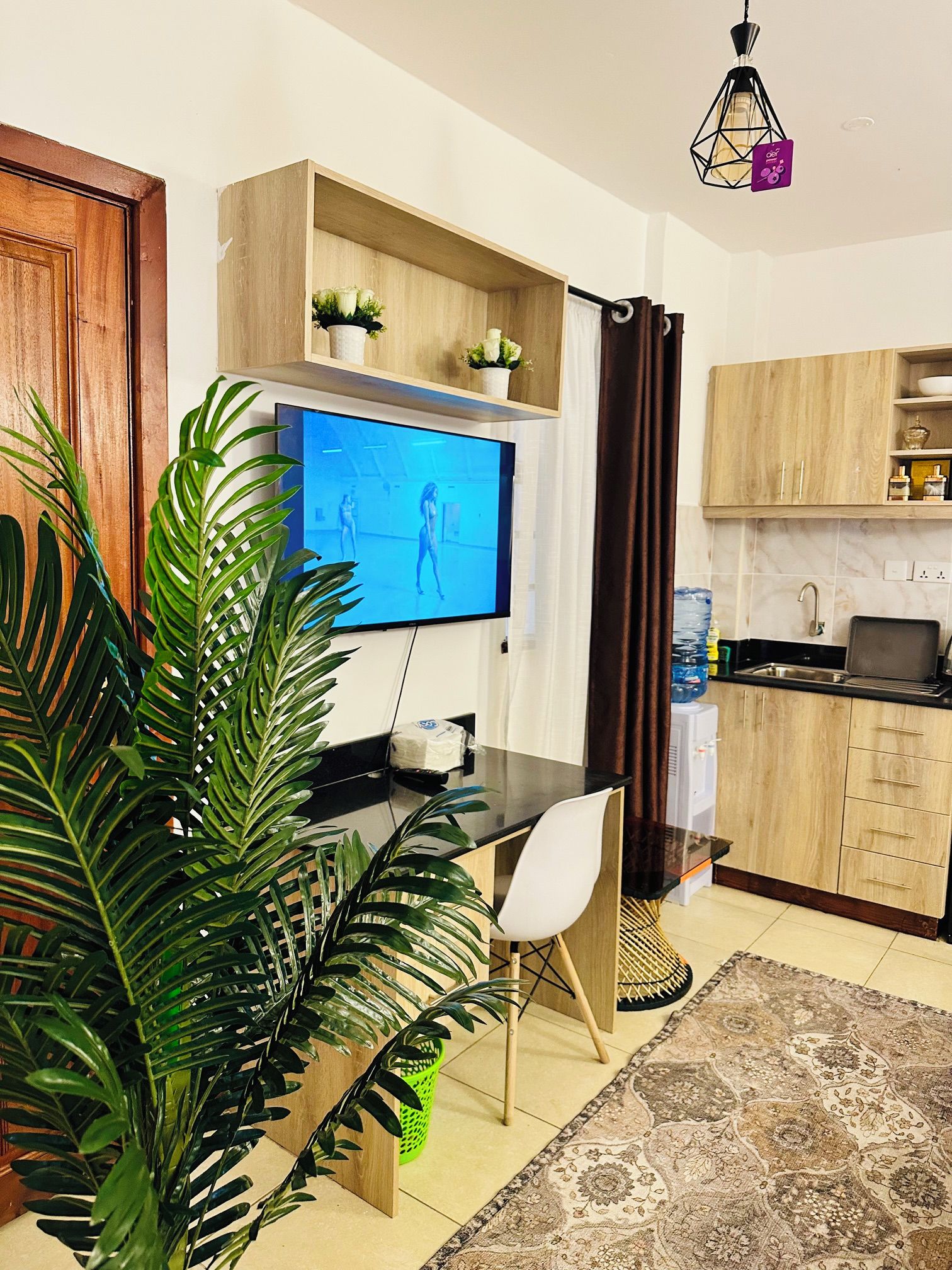Una sala de estar moderna con un televisor de pantalla plana, una mesa de comedor, una silla, una planta en maceta y una zona de cocina con armarios y dispensador de agua.