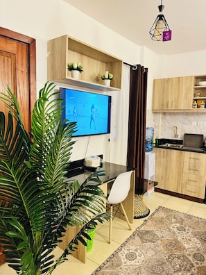 Una sala de estar moderna con un televisor de pantalla plana, una mesa de comedor, una silla, una planta en maceta y una zona de cocina con armarios y dispensador de agua.