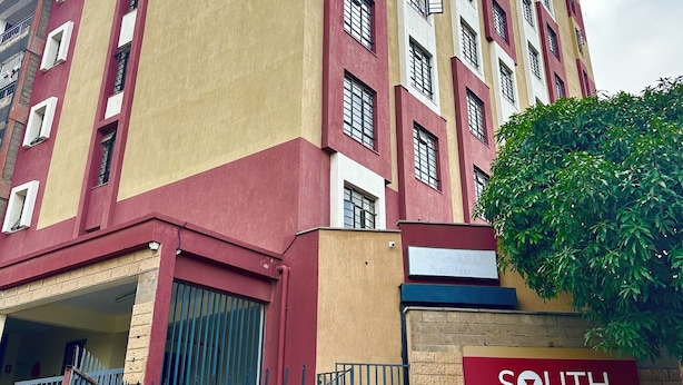 En bygning med flere etager, som har en rød og beige facade, en tydelig indgang med et skilt, og en grøn busk i forgrunden.