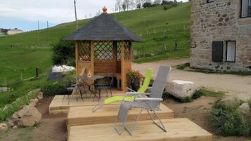 Un'area salotto all'aperto con un gazebo in legno, un edificio in pietra e una collina erbosa sullo sfondo.