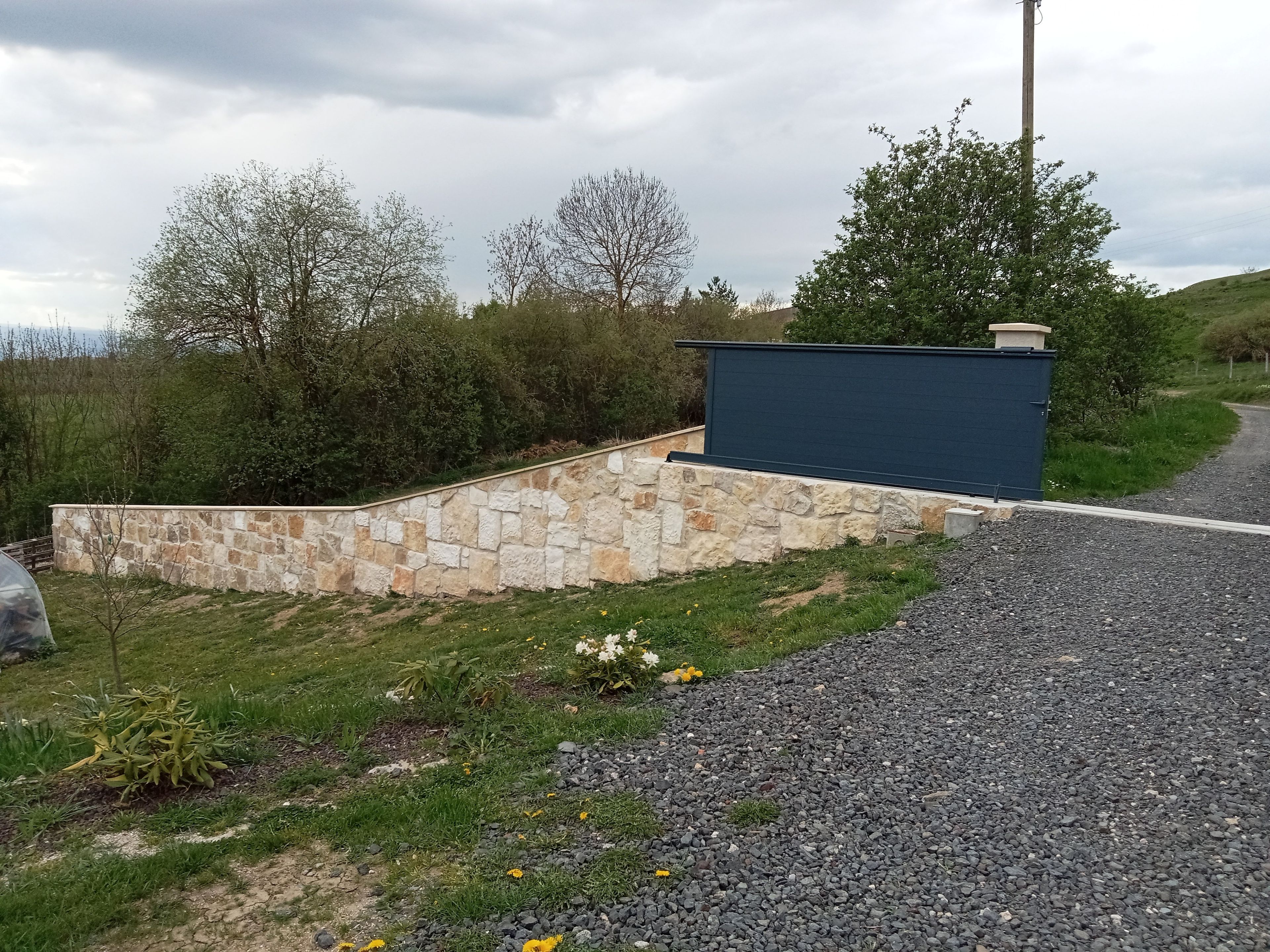 Un mur de pierre avec une structure bleue, un chemin de gravier et de la verdure.
