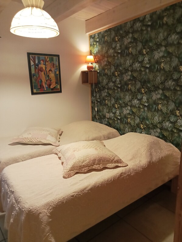 A room with two beds, a wall-mounted lamp, a framed painting, and a textured wallpaper.