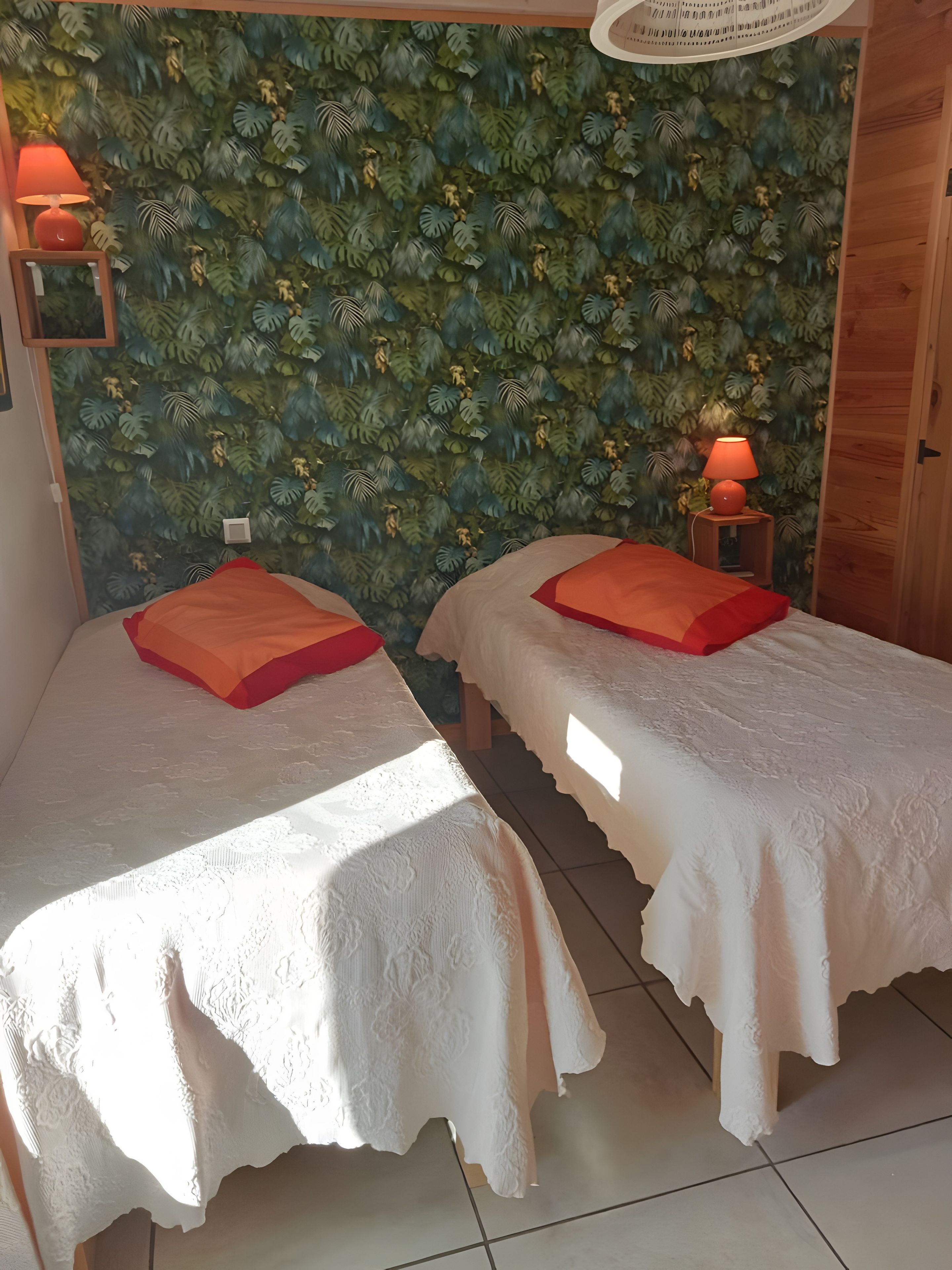 A room with two beds, each with a white bedspread and red pillows, against a wall with a leafy pattern. There are two wall-mounted lamps with red shades.