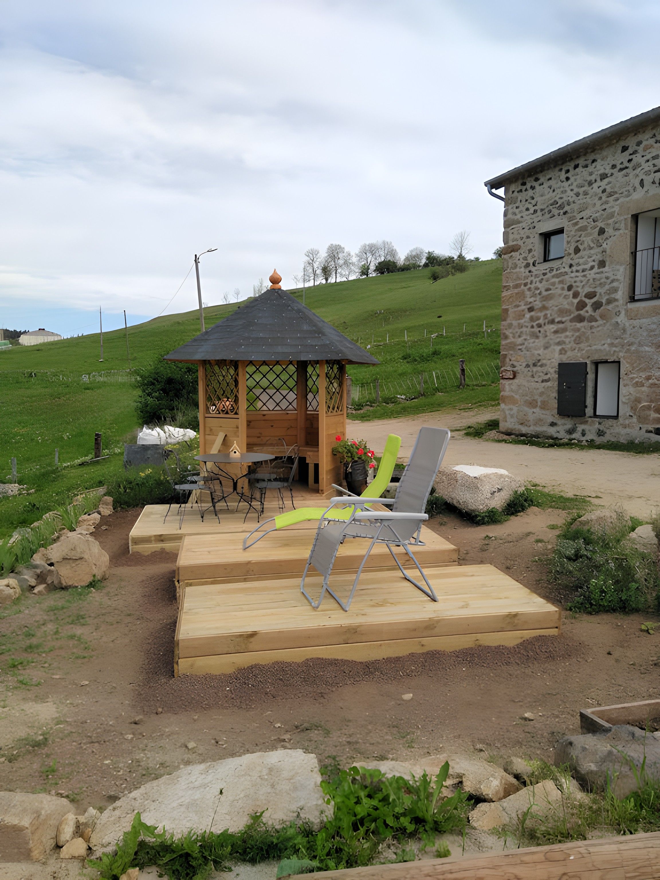 Un espace extérieur aménagé avec un kiosque en bois, un bâtiment en pierre et une colline verdoyante en arrière-plan.