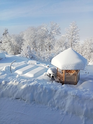 Une cabane en bois recouverte de neige, avec un toit également enneigé, entourée d’arbres chargés de neige et sous un ciel dégagé.
