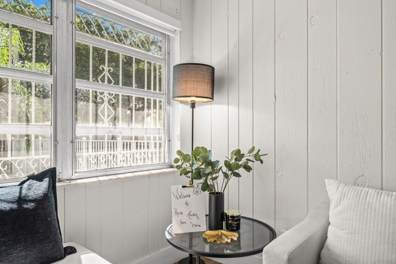A room with a white wooden wall, a window with a white railing, a black floor lamp, a small round table with a plant and a candle, and a black cushion on a white sofa.