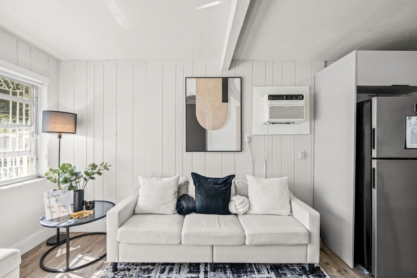 A modern living room with a white sofa, a glass coffee table, a black lamp, and a framed abstract artwork.