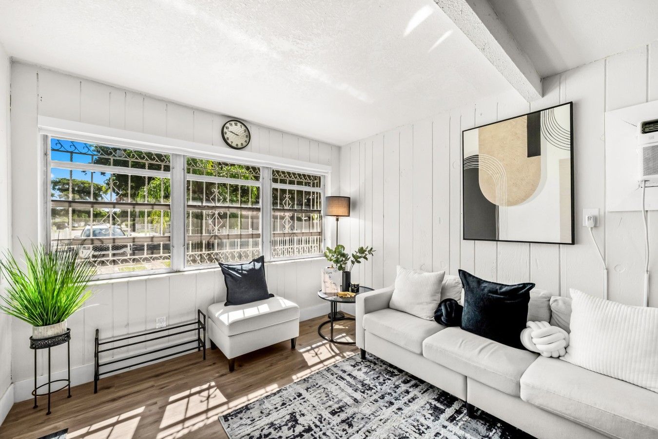 A modern living room with a white sofa, a coffee table, and a side table with a lamp. There is a large window with decorative iron bars and a clock above it.