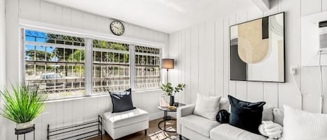 A modern living room with a white sofa, a coffee table, and a side table with a lamp. There is a large window with decorative iron bars and a clock above it.