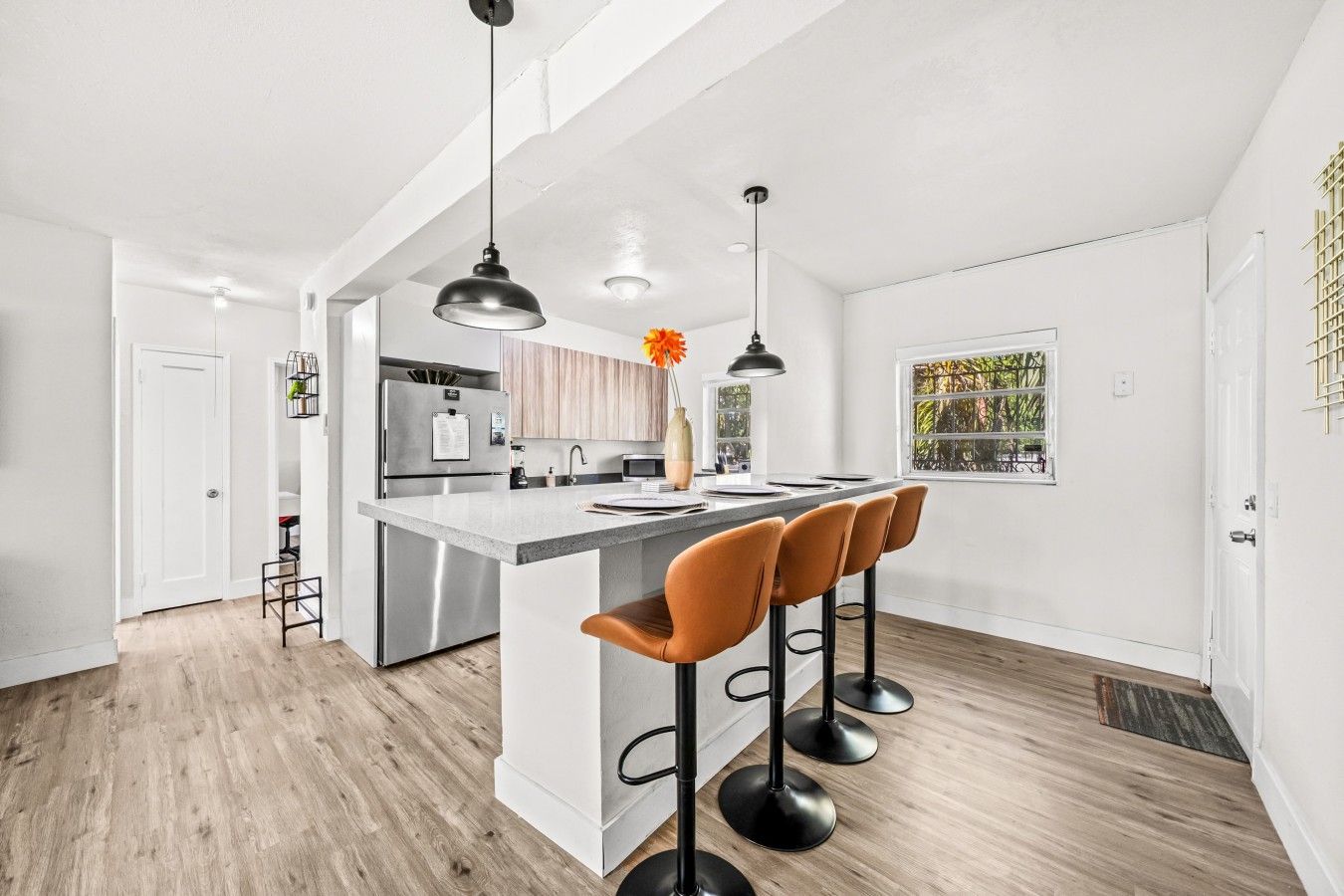 A modern kitchen with a central island, bar stools, pendant lighting, and a view of greenery outside.