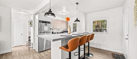 Cozinha moderna com ilha central, banquetas, iluminação pendente e vista para o verde lá fora.