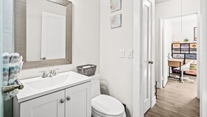 Een badkamer met een witte wastafel, een spiegel en een toilet. Op de wastafel staat een klein mandje en boven de wastafel een handdoekhouder met een handdoek.