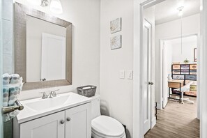 Une salle de bain avec un lavabo blanc, un miroir et des toilettes. On y trouve un petit panier sur le lavabo et un porte-serviettes avec une serviette.