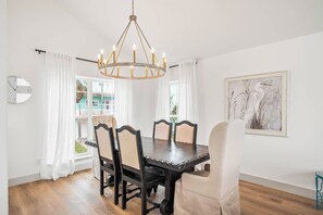 A dining area with a dark wooden table and chairs, a chandelier, and a painting on the wall. - Tan Lines & Fine Wines | Gorgeous Home + Walk to Beach! (Port Aransas)