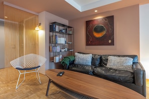 Ein modernes Wohnzimmer mit Ledersofa, Couchtisch aus Holz, einem Metallstuhl, einem Bücherregal und einem Bild an der Wand.