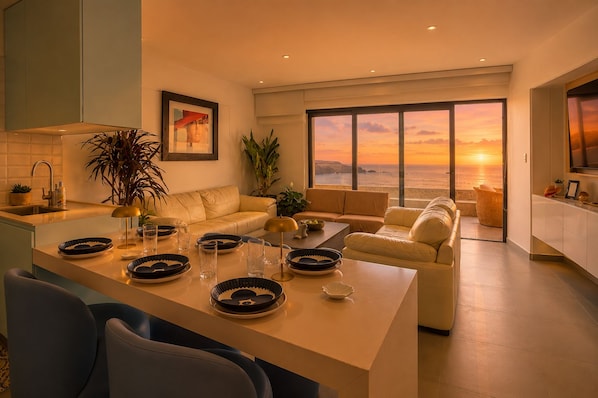 Ein modernes Wohnzimmer mit Küche, Essbereich und Sitzgelegenheit an einem großen Fenster mit Blick auf den Sonnenuntergang über dem Meer.