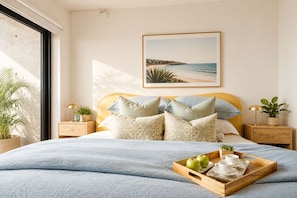 Ein Schlafzimmer mit einem Bett, Kissen, einem Tablett mit Äpfeln und Tassen sowie einem Gemälde vom Strand.