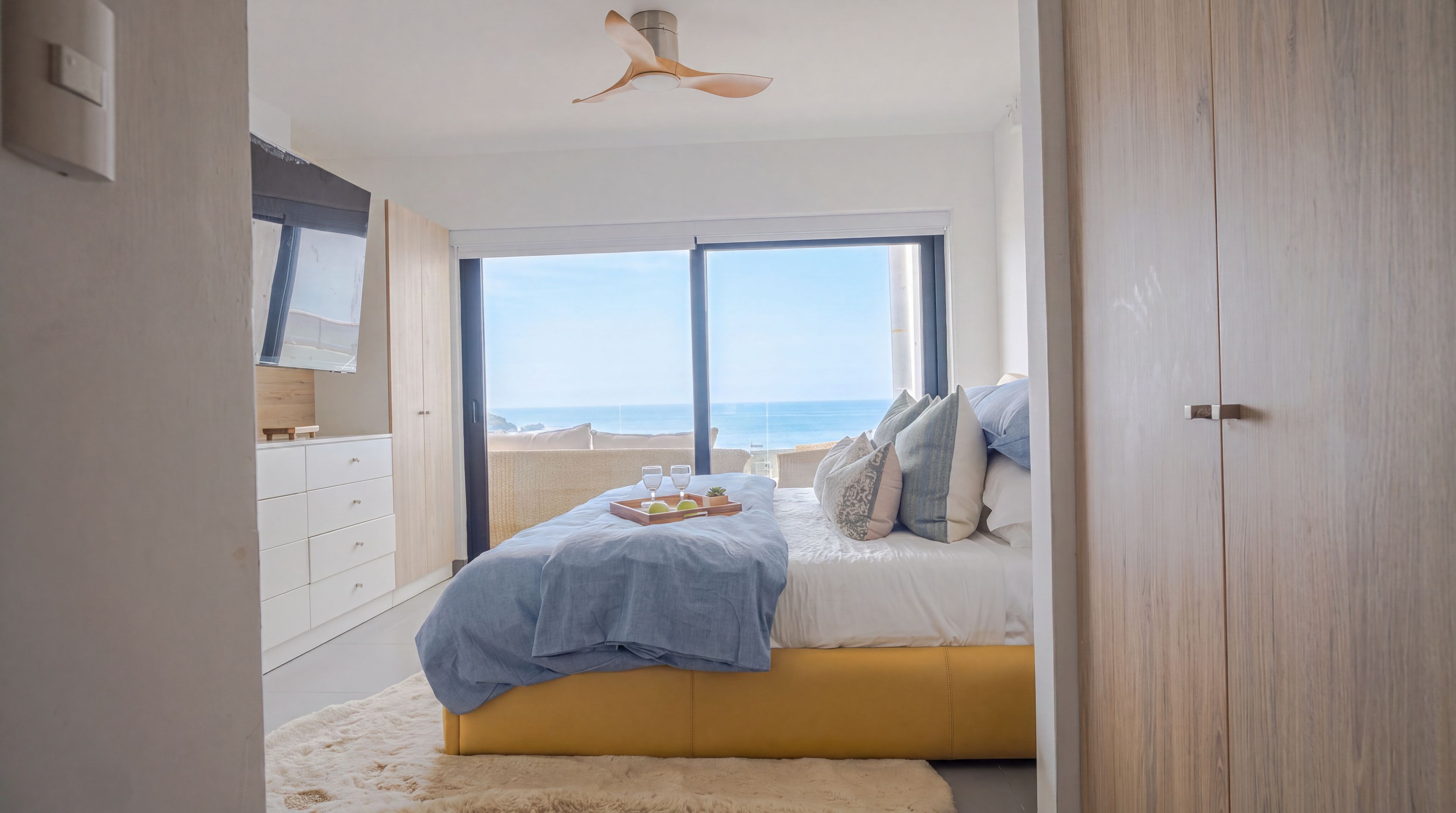 Un dormitorio con cama, mesita de noche, ventana con vista a la playa y ventilador de techo.