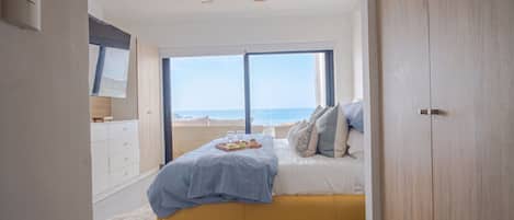 Quarto com cama, mesa de cabeceira, janela com vista para a praia e ventoinha no teto.