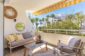 A balcony with a sofa, chairs, a coffee table, and a view of palm trees and buildings. - Marbella Playa Real | Pool & Sea View | 2 Bedrooms (Marbella)