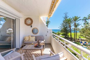 A balcony with a sofa, a table, and chairs, overlooking a pool and palm trees. - Marbella Playa Real | Pool & Sea View | 2 Bedrooms (Marbella)