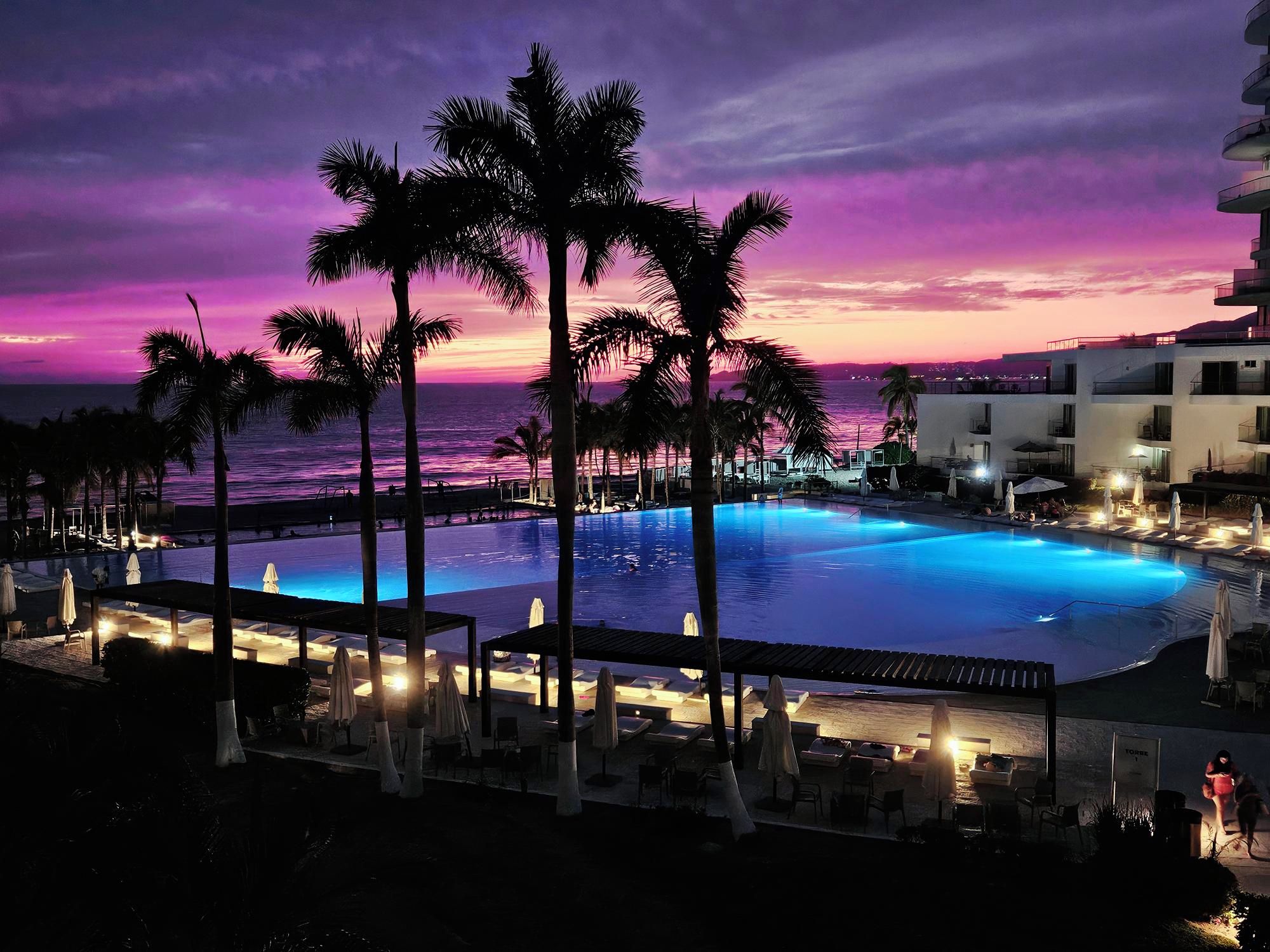 A pool area with lounge chairs and umbrellas, overlooking a body of water with a sunset sky.