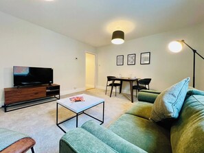 Ein Wohnzimmer mit einem Flachbildfernseher, einem Couchtisch, einer Couch, einem Esstisch mit Stühlen und einer Lampe.