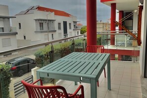 A balcony with a green table and red chairs, overlooking a street with parked cars and buildings.