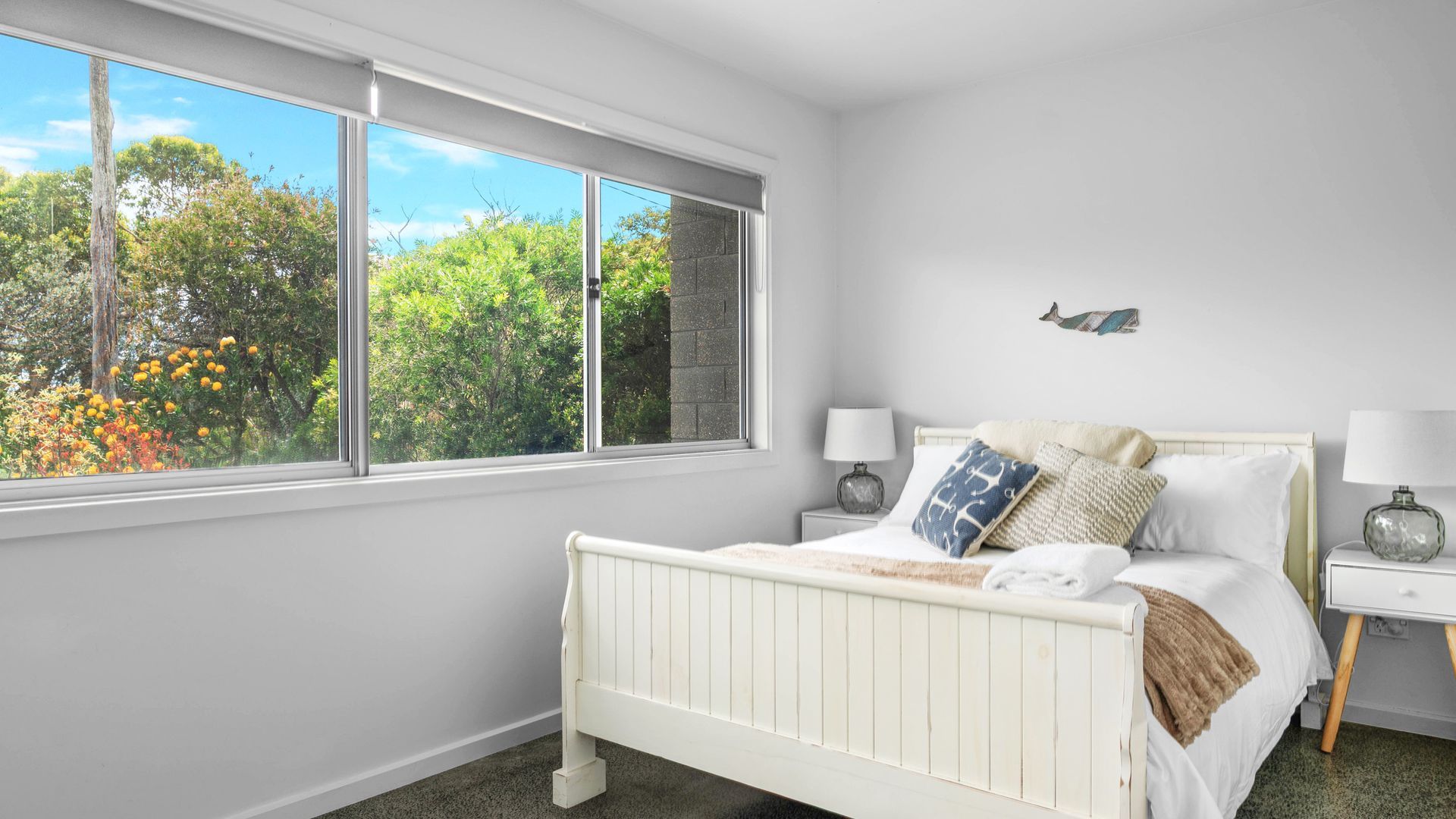 A bedroom with a white bed, two bedside tables, a window with a view of trees and a brick wall, and a small wall decoration.