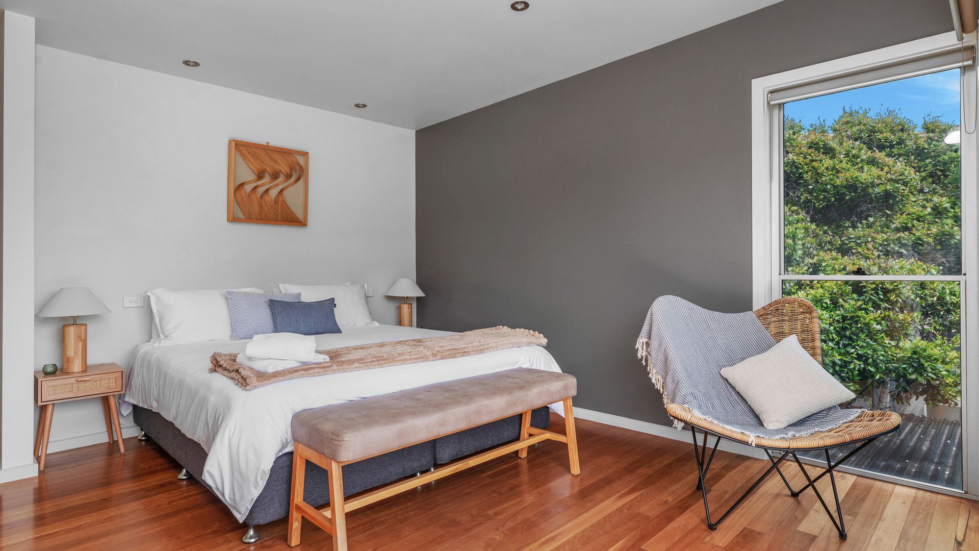 A modern bedroom with a bed, bench, bedside tables, and a view of greenery through a window.