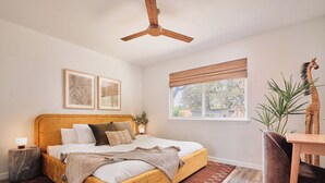 Ein Schlafzimmer mit einem Bett, einem Deckenventilator, einem Fenster mit Jalousien, einem Nachttisch, einer Lampe, einer Pflanze und einer Giraffefigur.