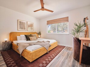 A bedroom with a bed, a ceiling fan, a window with blinds, a nightstand, a lamp, a plant, and a giraffe statue. - Casa Peña | Designer 3BR Retreat w/ Fire Pit (Austin)